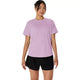 En person står med ansigtet vendt fremad iført en Asics Core Ss Top, Dame i farven Hazy Lilac med refleksdetaljer og sorte shorts, foran en ensfarvet hvid baggrund.