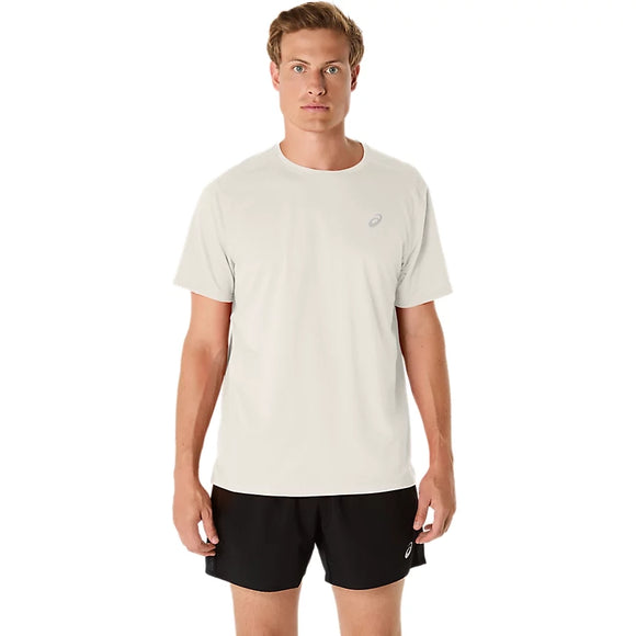 En person står med ansigtet vendt fremad iført en ASICS Core Ss Top, Herre, Cream – en kortærmet løbetrøje til mænd i cremefarve – og sorte shorts med et lille logo på hvert stykke tøj, foran en ensfarvet hvid baggrund.