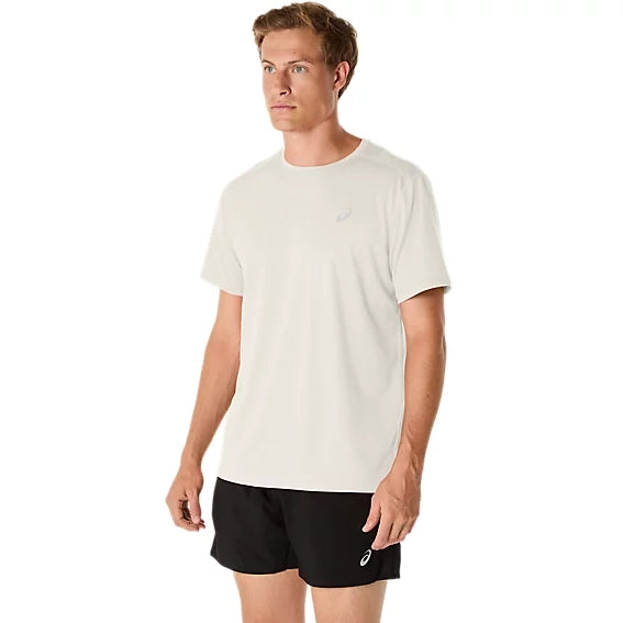 En mand står foran en ensfarvet hvid baggrund iført en ASICS Core SS Top, Herre, i farven creme samt sorte sportsshorts. Begge beklædningsgenstande er forsynet med små ASICS-logoer. Han kigger let til siden med et neutralt udtryk i ansigtet.