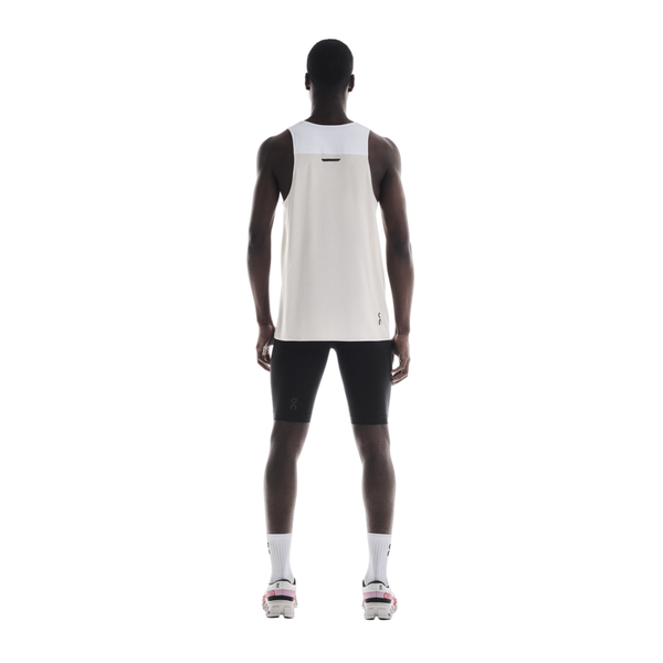 En person står med ansigtet vendt mod kameraet iført On Performance Tank, Herre, White/Silver, sorte shorts, hvide sokker og hvide sportssko med pink detaljer på en almindelig hvid baggrund.