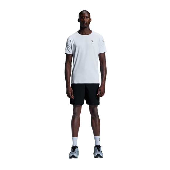 En mand kigger fremad mod en almindelig hvid baggrund iført On Performance-T, Herre i hvid/sølv, sammen med sorte shorts, hvide sokker og grå sportssko.