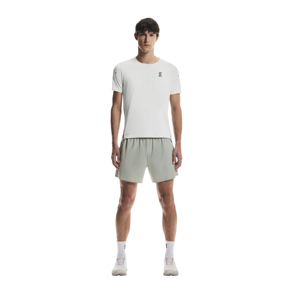 En mand ser frem for sig iført On Performance-T, Herre, Iceberg/Tin, lysegrønne shorts, hvide strømper og hvide sneakers på en almindelig hvid baggrund.