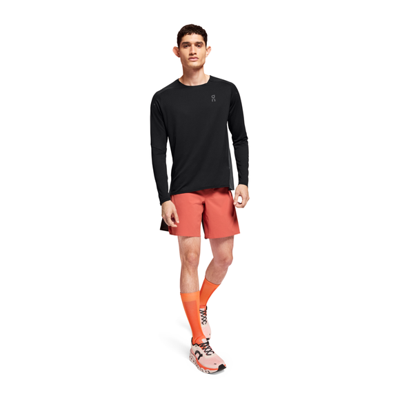En mand med kort mørkt hår og et neutralt udtryk står med ansigtet fremad i en On Performance Long-T, Herre, Black/Eclipse, parret med koralfarvede sportsshorts, orange knæstrømper og pink løbesko med orange snørebånd.