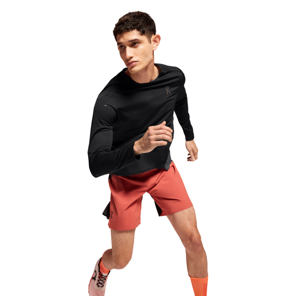 En mand i en On Performance Long-T, Herre, Black/Eclipse, med røde shorts, orange sokker og hvide løbesko ses løbe eller posere i en løbestilling mod en hvid baggrund.
