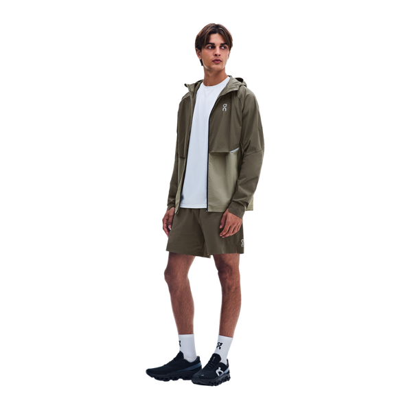 En mand er iført en tofarvet olivengrøn On Core-jakke, Herre, Bark/Vetiver, en hvid T-shirt, olivengrønne shorts, hvide sokker og sorte sportssko.