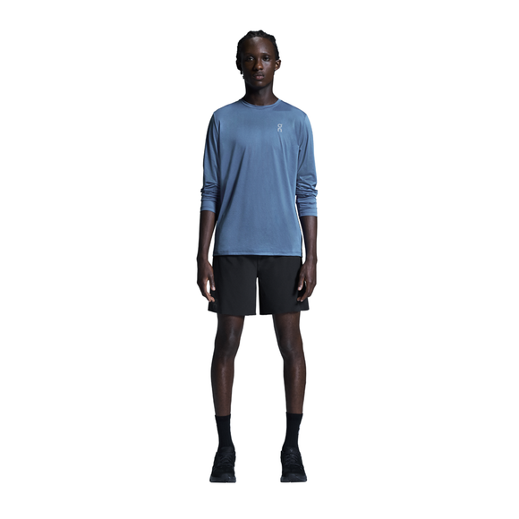 En person står med ansigtet vendt fremad foran en ensfarvet hvid baggrund, iført en blå langærmet skjorte, ON 7 Performance Shorts, Herre, Sort, sorte sokker og sorte sneakers. Armene hviler naturligt langs siden, hvilket fremhæver den perfekte kombination af komfort og stil, som disse letvægts-løbeshorts fra ON byder på.