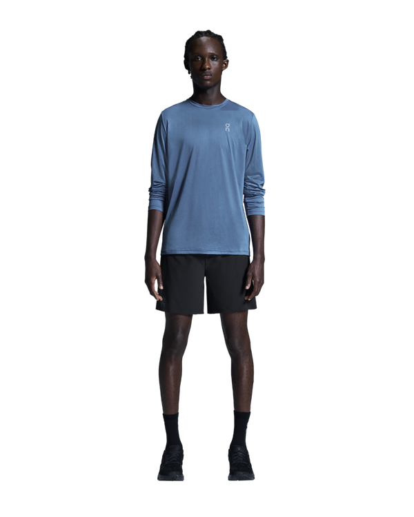 En person står med ansigtet vendt fremad foran en ensfarvet hvid baggrund, iført en blå langærmet skjorte, ON 7 Performance Shorts, Herre, Sort, sorte sokker og sorte sneakers. Armene hviler naturligt langs siden, hvilket fremhæver den perfekte kombination af komfort og stil, som disse letvægts-løbeshorts fra ON byder på.