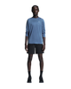 En person står med ansigtet vendt fremad foran en ensfarvet hvid baggrund, iført en blå langærmet skjorte, ON 7 Performance Shorts, Herre, Sort, sorte sokker og sorte sneakers. Armene hviler naturligt langs siden, hvilket fremhæver den perfekte kombination af komfort og stil, som disse letvægts-løbeshorts fra ON byder på.
