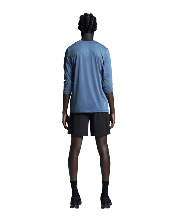 En person står med ryggen til kameraet og er iført en blå langærmet skjorte og et par ON 7 Performance Shorts, Herre, Sort – perfekte letvægts løbeshorts til enhver aktivitet. Sorte sokker og sorte sneakers fuldender looket, der fremstår på en ensfarvet hvid baggrund. Personens arme hænger naturligt ned langs siden.