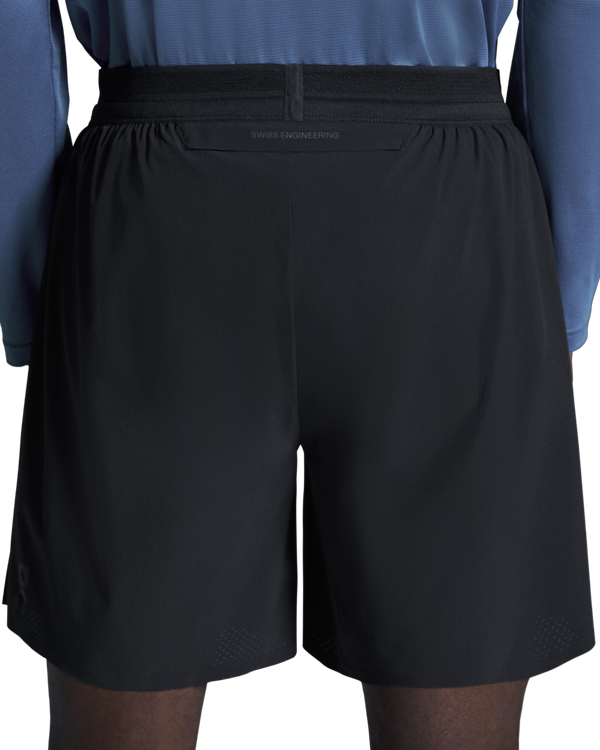 Et billede bagfra af en person, der bærer ON 7 Performance Shorts, Herre, Sort og en blå langærmet trøje. På linningen på de lette løbeshorts står der »SWISS ENGINEERING« med små bogstaver.