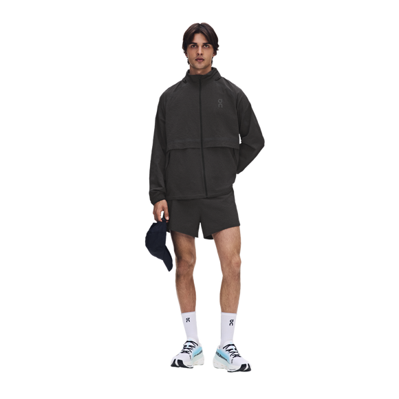 En mand står med ansigtet vendt fremad iført On Performance Volt-jakken til mænd i farven Espresso, kombineret med matchende shorts, hvide sokker med sorte logoer og hvide sneakers. Han holder en mørk kasket i højre hånd foran en ensfarvet hvid baggrund.