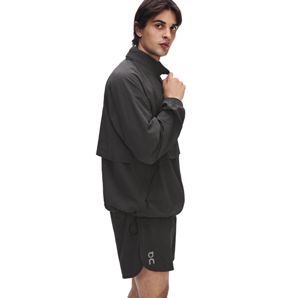 En person iført ON Performance Volt-jakken (herre) i farven Espresso og tilhørende shorts står foran en hvid baggrund og kigger over skulderen. Shortsene har et lille ON-logo tæt på sømmen.