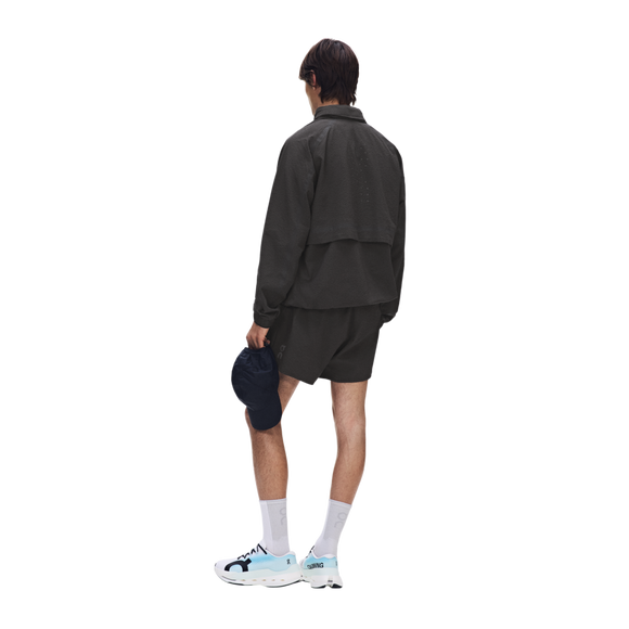 En person står med ryggen til og er iført en ON Performance Volt-jakke til mænd i farven Espresso, matchende shorts, hvide sokker og hvide sneakers med blå detaljer. Personen holder en mørk kasket i den ene hånd foran en ensfarvet hvid baggrund.