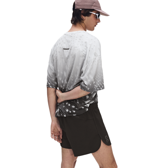En person iført en lysegrå og hvidmønstret skjorte, ON Performance Volt Shorts Herre Espresso, en lyserød kasket og solbriller står foran en ensfarvet hvid baggrund med ryggen til og den ene hånd bag ryggen.