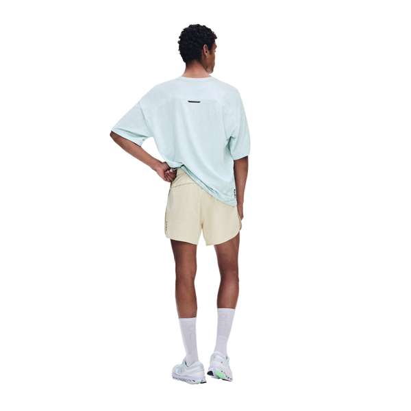 En person ses bagfra iført en lyseblå oversized T-shirt og ON Performance Volt Shorts, Herre, Hare – vandafvisende løbeshorts – kombineret med hvide sokker og sneakers foran en ensfarvet hvid baggrund.