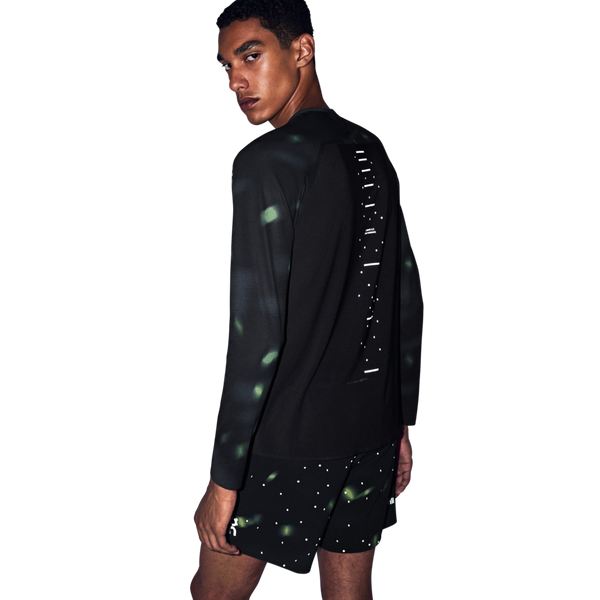 En mand iført en sort Performance Long-T Lumos 3, Herre med reflekterende grønne detaljer og en lodret række prikker på ryggen, kombineret med sorte shorts med et hvidt prikmønster.