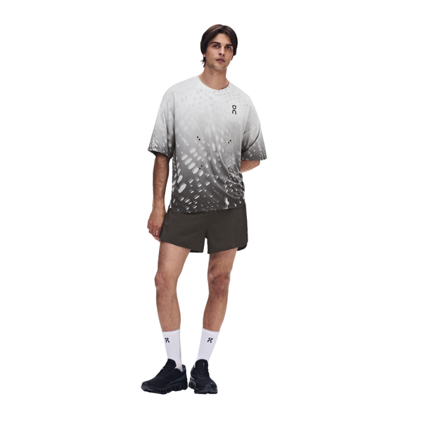 En mand står foran en ensfarvet baggrund iført en ON Performance Volt-T, Herre, Espresso, mørke shorts, hvide strømper og sorte sneakers. Den hurtigtørrende trøje sikrer komfort. Han har den ene hånd bag ryggen, mens han kigger lige frem.