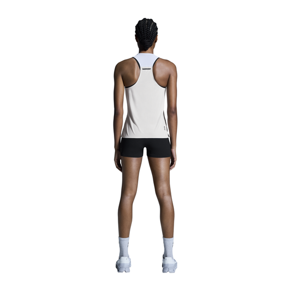 En person med flettet hår står med ansigtet vendt mod kameraet iført On Performance Tank, Dame i hvid/sølv, sorte shorts, hvide sokker og hvide sportssko på en almindelig hvid baggrund.