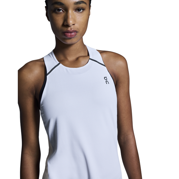 En kvinde bærer On Performance Tank, Dame, White/Silver - en hvid sportstop uden ærmer med sort kant og et lille logo på venstre bryst - mens hun står på en almindelig hvid baggrund og kigger fremad.