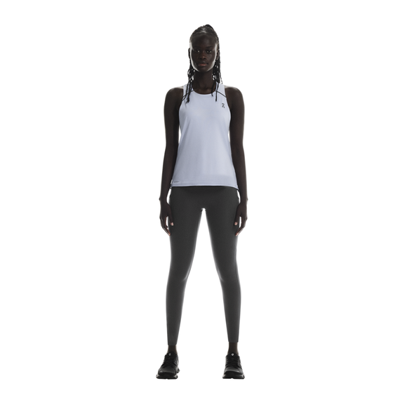 En person med lange fletninger står og kigger fremad, iført On Performance Tank til kvinder i Nimbus/Lilac, sammen med mørkegrå leggings og sorte sneakers på en almindelig hvid baggrund.