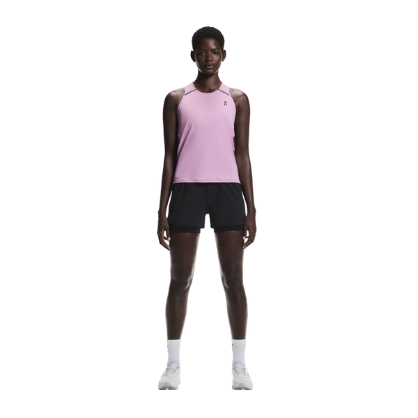 En person står og kigger fremad iført On Performance Tank, Dame i Sakura/Dewberry med sorte shorts, hvide strømper og hvide sneakers på en hvid baggrund - perfekt åndbar løbetøj til aktive dage.