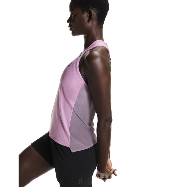 En person i On Performance Tank til kvinder i Sakura/Dewberry og sorte shorts står sidelæns og holder den ene fod bag sig i en quadriceps-strækning mod en almindelig hvid baggrund.