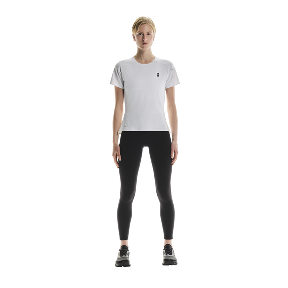 En person står med ansigtet vendt fremad iført en ON Performance-T, Dame, hvid/sølv, kombineret med sorte leggings og sorte sportssko, foran en ensfarvet hvid baggrund.