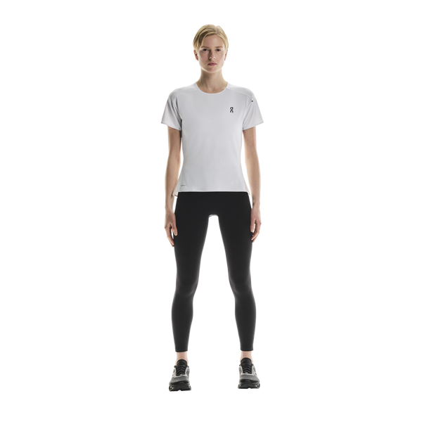 En person står med ansigtet vendt fremad iført en ON Performance-T, Dame, hvid/sølv, kombineret med sorte leggings og sorte sportssko, foran en ensfarvet hvid baggrund.