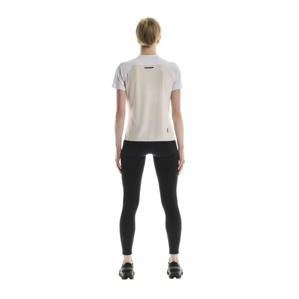 En person står med ryggen til kameraet iført en ON Performance-T, Dame, hvid/sølv, kombineret med sorte leggings og sorte sportssko foran en ensfarvet hvid baggrund.