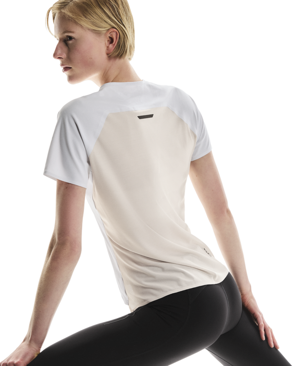 Der ses en kvinde med kort blondt hår set bagfra, mens hun strækker sig foran en hvid baggrund. Hun er iført ON Performance-T, Dame, hvid/sølv, samt sorte leggings.