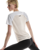 Der ses en kvinde med kort blondt hår set bagfra, mens hun strækker sig foran en hvid baggrund. Hun er iført ON Performance-T, Dame, hvid/sølv, samt sorte leggings.