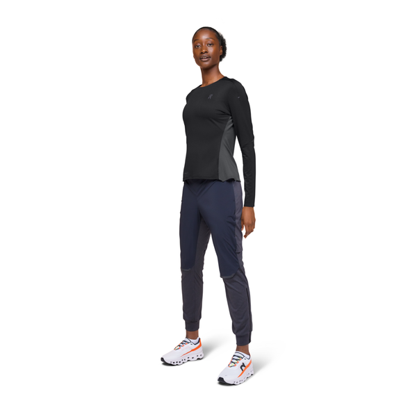 En kvinde kigger fremad iført On Performance Long-T, Dame, Black/Eclipse, sammen med mørke joggings og hvide løbesko med orange detaljer - ideel træningstøj dame - mod en almindelig hvid baggrund.