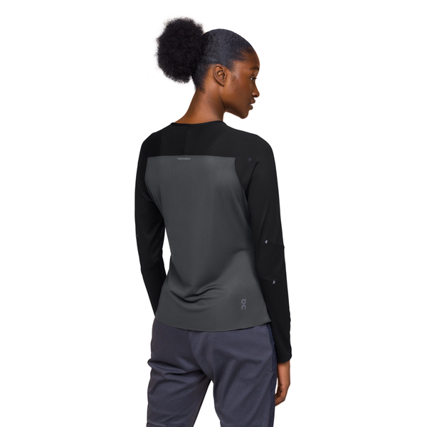 En kvinde med mørkt, tilbagestrøget hår står og kigger væk, iført On Performance Long-T, Dame, Black/Eclipse top og mørke bukser. Baggrunden er helt hvid.
