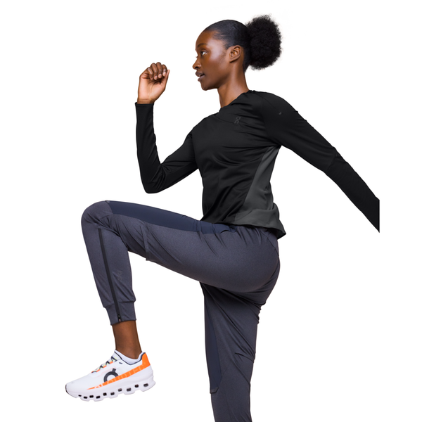 En kvinde i On's Performance Long-T, Dame, Black/Eclipse laver høje knæ i grå joggingbukser og hvide sneakers med orange detaljer på en almindelig hvid baggrund.