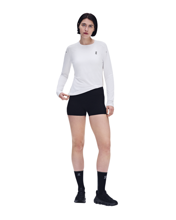 En person står foran en hvid baggrund iført en On Performance Long-T, Dame, hvid/sølv, sammen med sorte shorts, sorte sokker og sorte løbesko. For at demonstrere fordelene ved åndbart løbetøj kigger vedkommende fremad, mens vedkommende holder fast i trøjens nederste kant.