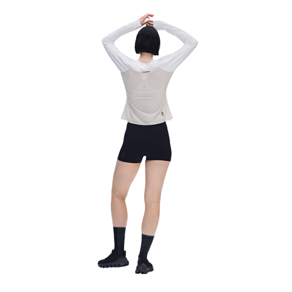En person står med ryggen til, armene løftet og hænderne samlet over hovedet. Personen har en On Performance Long-T, Dame i hvid/sølv på, kombineret med sorte shorts, sorte strømper og sorte sportssko. Baggrunden er helt hvid.