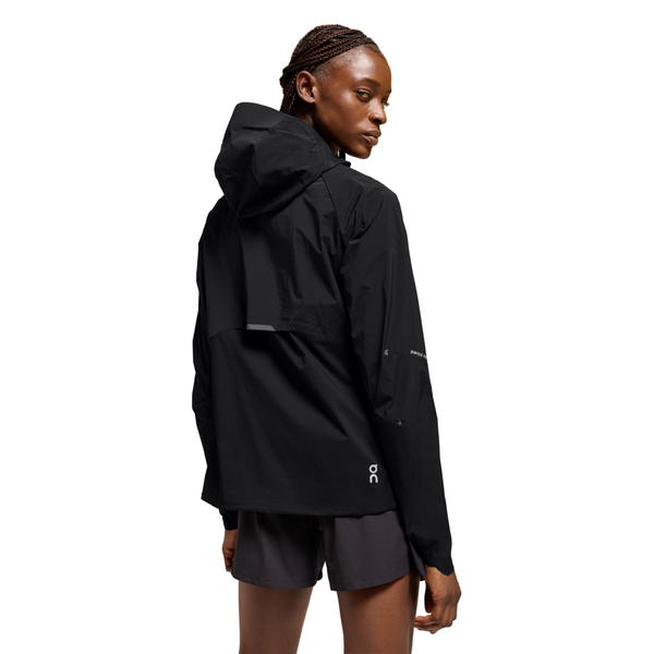 En person iført On Core Jacket til kvinder i sort og mørke shorts står med ryggen til og drejer hovedet lidt til siden mod en almindelig hvid baggrund.