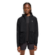En kvinde med lange fletninger og et neutralt udtryk står med ansigtet vendt fremad iført On Core Jacket, Dame, Black over en sort skjorte og shorts. Den vindafvisende jakke har et lille logo på venstre bryst.