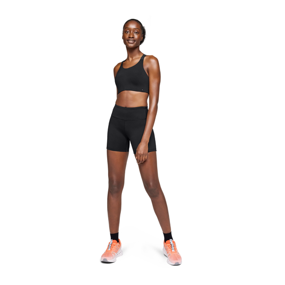 En kvinde står med front mod en almindelig hvid baggrund iført On Performance Tights Short, Dame, sort sports-bh, sorte sokker og orange sneakers.