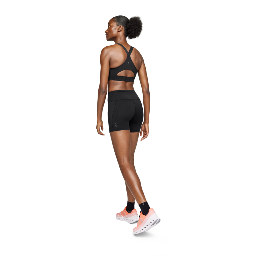 En kvinde står på en hvid baggrund iført On Performance Tights Short, Dame, Black med sorte sokker og lyserøde sneakers. Hun kigger væk og drejer hovedet til venstre for at fremhæve det stilfulde design.
