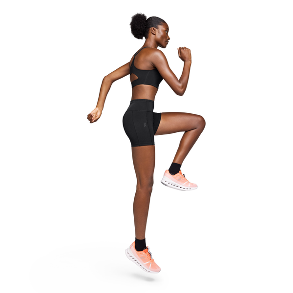 En kvinde iført On Performance Tights Short, Dame, Black og pink løbesko laver en høj knæøvelse sidelæns mod en hvid baggrund med venstre knæ løftet og højre arm bøjet fremad.