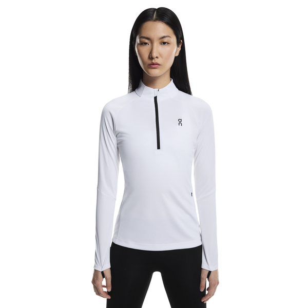 En kvinde med langt glat hår bærer On Climate Shirt, Dame, White - en tætsiddende, langærmet skjorte med lynlås foran og sort logo, fremstillet af hurtigtørrende, åndbart stof - sammen med sorte bukser på en almindelig hvid baggrund.