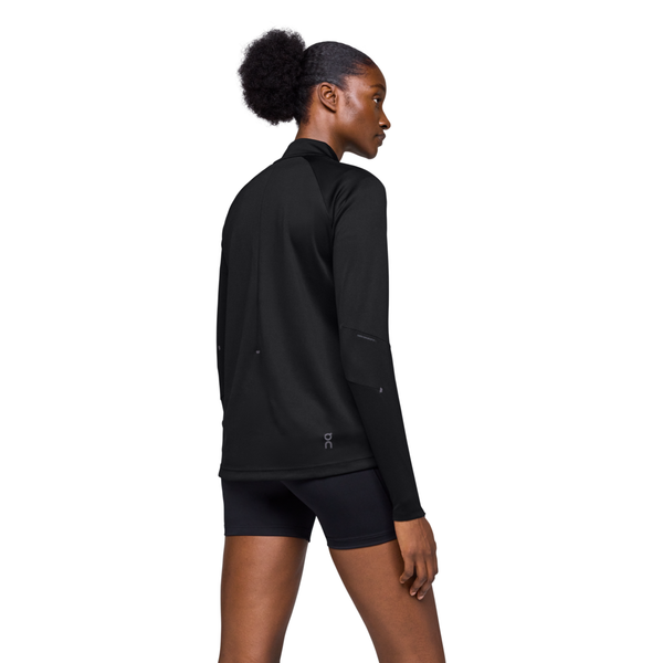 En kvinde med mørkt hår i en pjuske bærer On Climate Shirt, Dame, Black med lange ærmer og sorte shorts, mens hun står og kigger til siden mod en almindelig hvid baggrund.