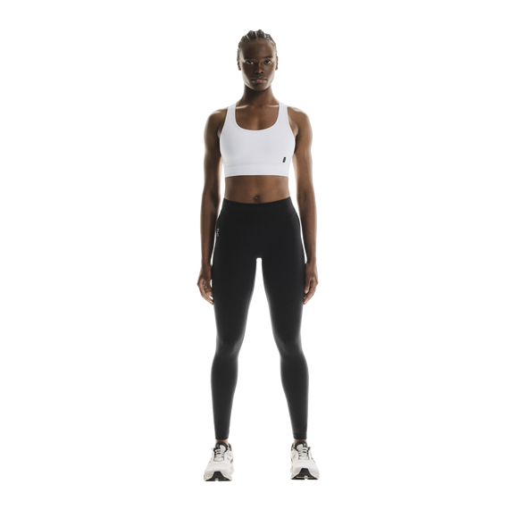 En kvinde står med ansigtet fremad iført ON Core Bra, Dame, hvid, samt sorte leggings og hvide sportssko foran en ensfarvet hvid baggrund, klar til træning med lav intensitet.