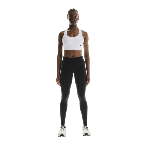 En kvinde står med ansigtet fremad iført ON Core Bra, Dame, hvid, samt sorte leggings og hvide sportssko foran en ensfarvet hvid baggrund, klar til træning med lav intensitet.