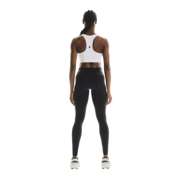 En person står med ryggen til kameraet og har ON Core Bra på, modellen »Dame« i hvidt, kombineret med sorte leggings og hvide sneakers foran en ensfarvet hvid baggrund – det perfekte tøj til træning med lav intensitet.