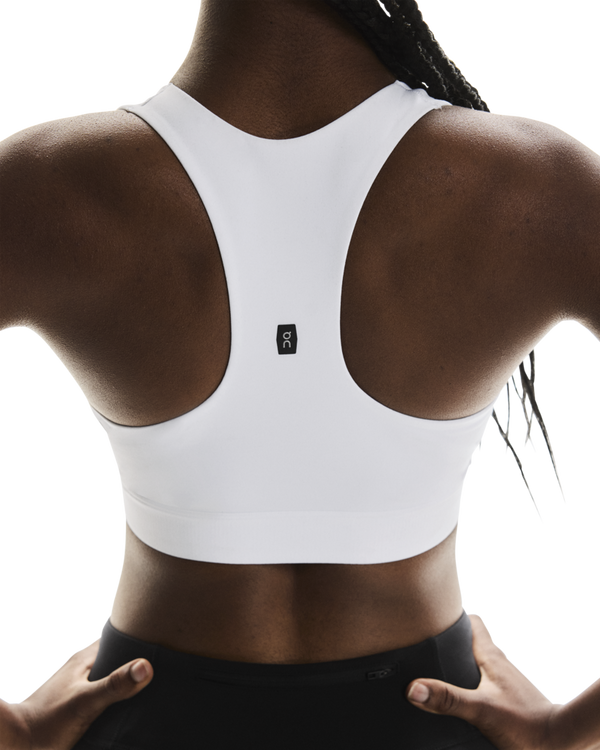 En person med mørk hud iført ON Core Bra, Dame, White — en behagelig sports-bh i racerback-design — samt sorte underdele, set bagfra. Sports-bh'en har et lille sort-hvidt ON-logo midt øverst på ryggen. Hænderne hviler på hofterne – et ideelt look til træning med lav intensitet.