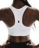 En person med mørk hud iført ON Core Bra, Dame, White — en behagelig sports-bh i racerback-design — samt sorte underdele, set bagfra. Sports-bh'en har et lille sort-hvidt ON-logo midt øverst på ryggen. Hænderne hviler på hofterne – et ideelt look til træning med lav intensitet.