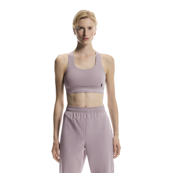 En person står og kigger fremad iført On Core Bra, Dame, Heron - en behagelig, lyslilla sports-bh sammen med matchende joggingbukser - på en almindelig hvid baggrund, perfekt til løb.