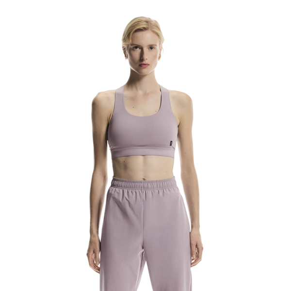 En person står og kigger fremad iført On Core Bra, Dame, Heron - en behagelig, lyslilla sports-bh sammen med matchende joggingbukser - på en almindelig hvid baggrund, perfekt til løb.